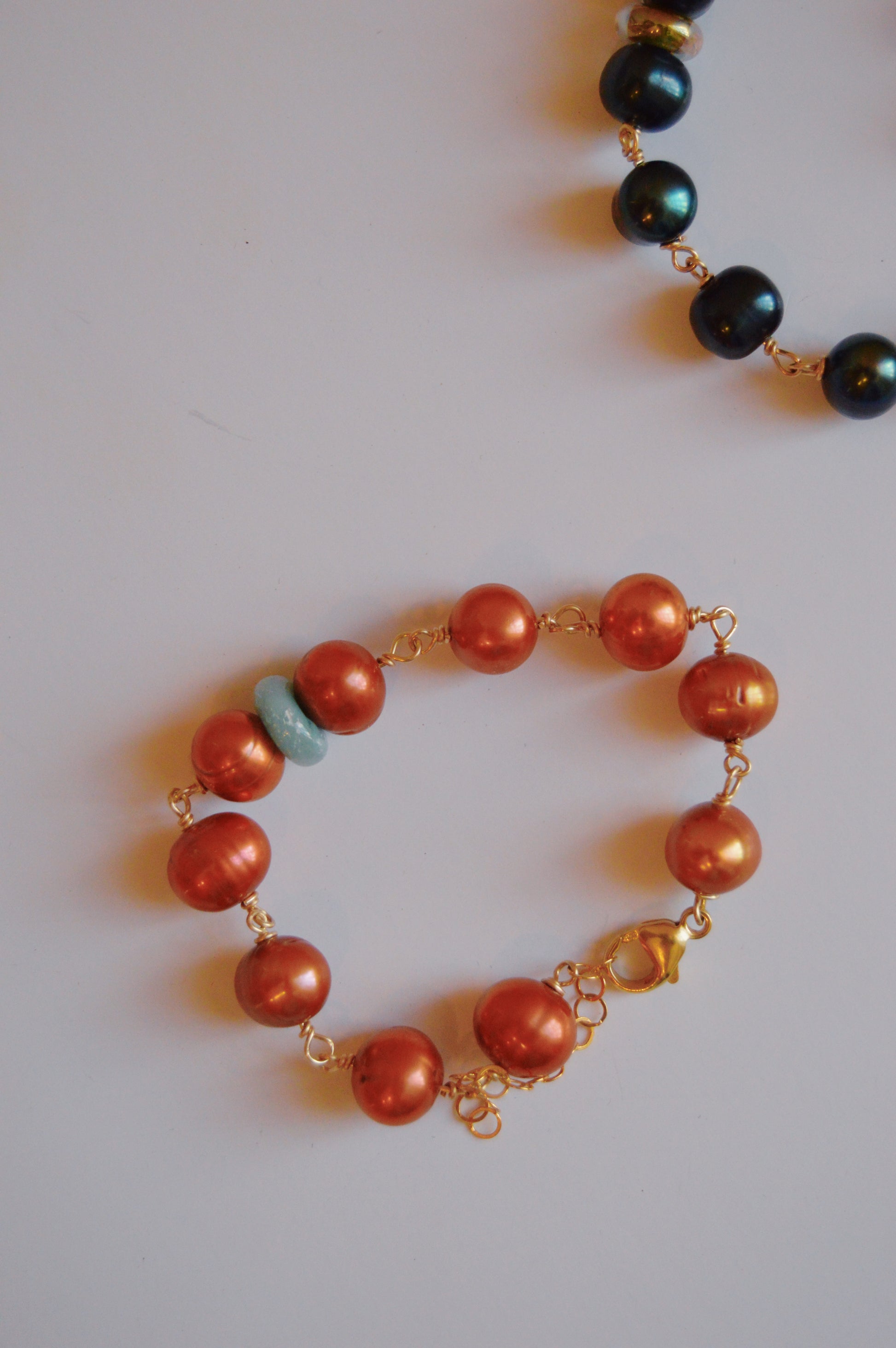 The Pearl and Glass Bracelet in copper is shown on a white background, featuring large handmade lampwork beads and gold-filled wire links. At the top, a portion of another bracelet with dark beads is visible.