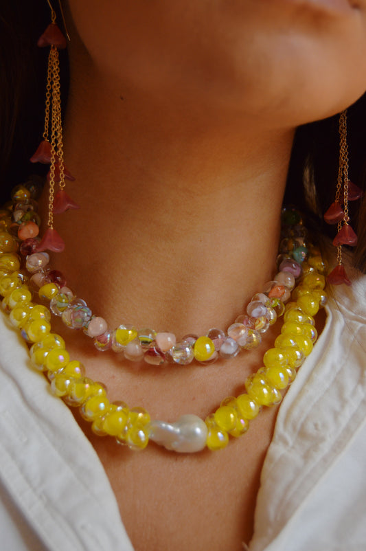 A woman wearing Stack #6—two beaded necklaces (yellow, clear & pastel beads) and long gold earrings with pink floral charms, styled with a white top. Enjoy 20% off on similar styles during our giveaway!.