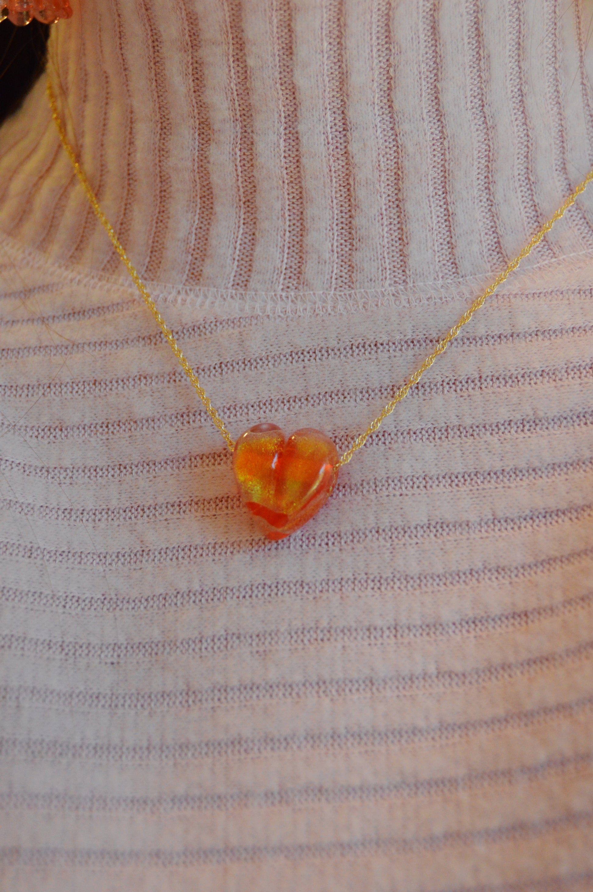 A person wears a ribbed light pink turtleneck sweater paired with a delicate gold-filled chain necklace featuring the Glass Heart Necklaces- Reds & Oranges, showcasing a small handmade lampwork glass heart pendant.