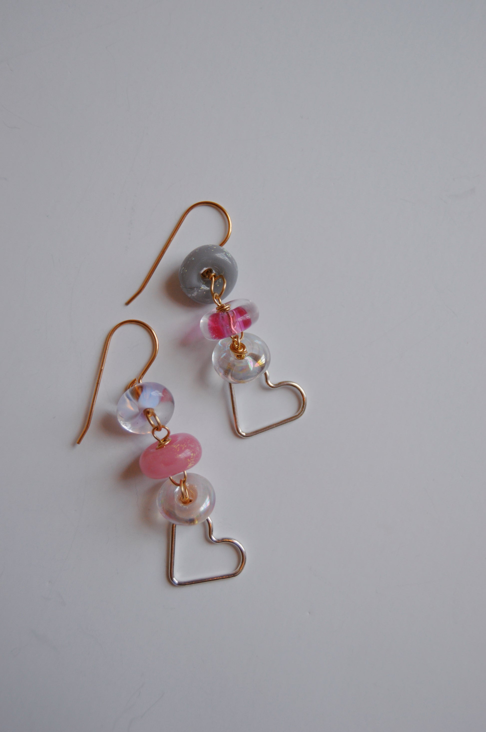 Silver Heart Drops: Elegant earrings with gold hooks, stacked handmade lampwork glass beads in gray, pink, and clear, each finished with a gold wire heart-shaped charm. Handcrafted jewelry displayed on a plain white surface.