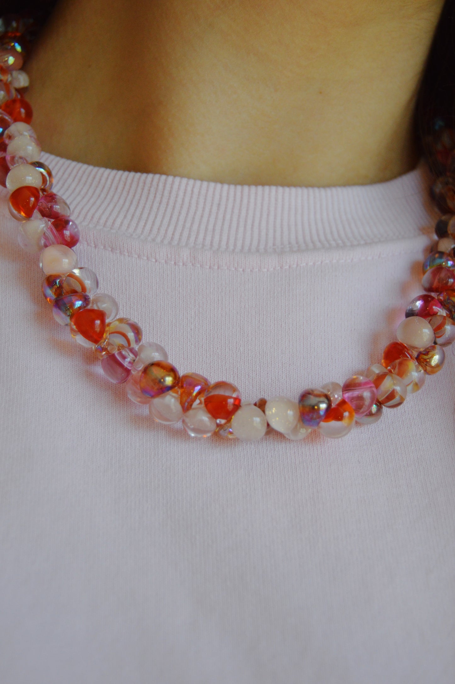 A person wears the Valentine Bubble Glass Necklace- Lovebug, a colorful handmade piece featuring red, pink, and iridescent round beads, resting at the base of their neck with a light pink shirt.