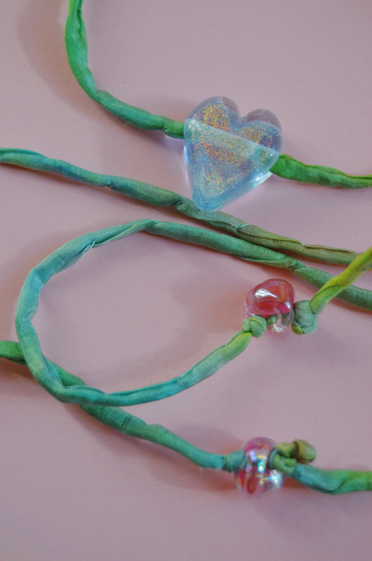 The Silk Glass Heart- Blue, Green, & Pink features handcrafted jewelry with blue-green silk cords, a large iridescent heart bead, and two small pink lampwork glass beads, displayed on a light pink background.
