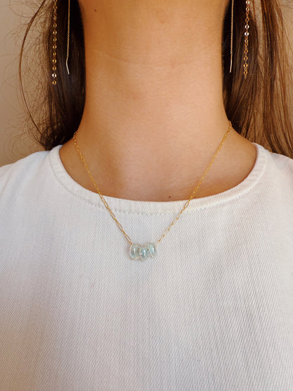 A person wearing a white top showcases the Dainty Donut Layer—a 14K gold chain necklace with light blue glass donut beads and a gold-filled clasp—paired with long dangling gold earrings, highlighting the jewelry and neckline.