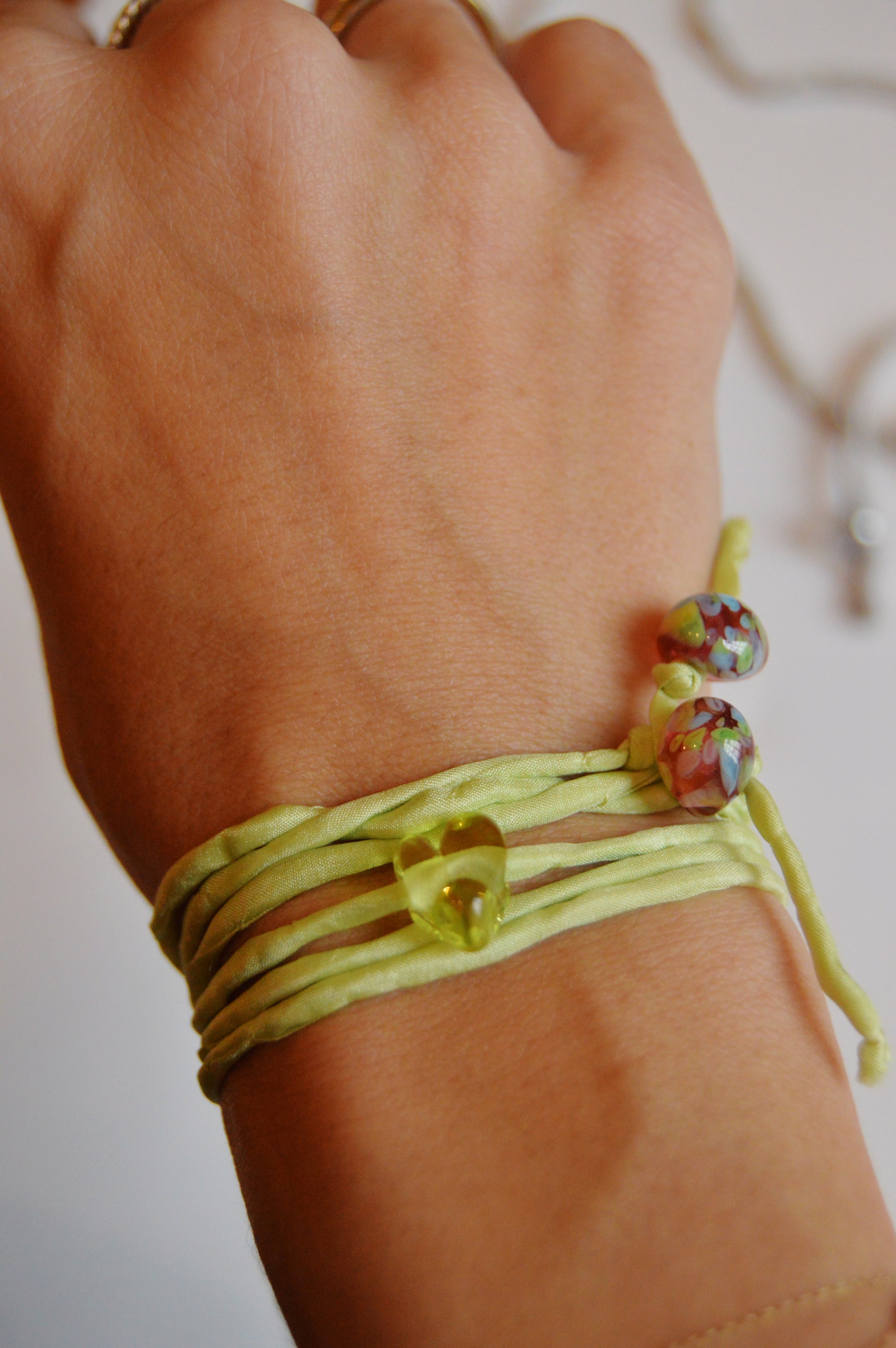 A hand wears the Mini Silk Glass Heart- Light Green wrap bracelet, featuring a small yellow heart bead, two multicolored round beads, and handmade lampwork glass accents. Multiple light green silk strands elegantly wrap the wrist.
