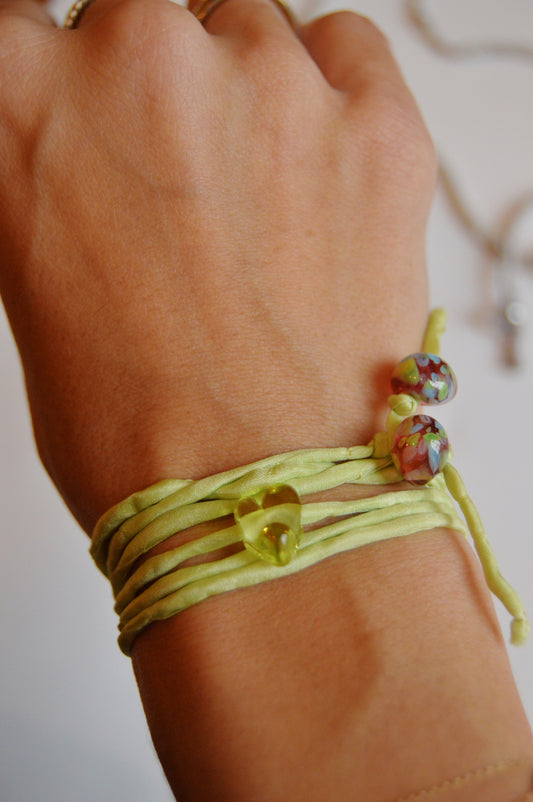 A hand wears the Mini Silk Glass Heart- Light Green wrap bracelet, featuring a small yellow heart bead, two multicolored round beads, and handmade lampwork glass accents. Multiple light green silk strands elegantly wrap the wrist.