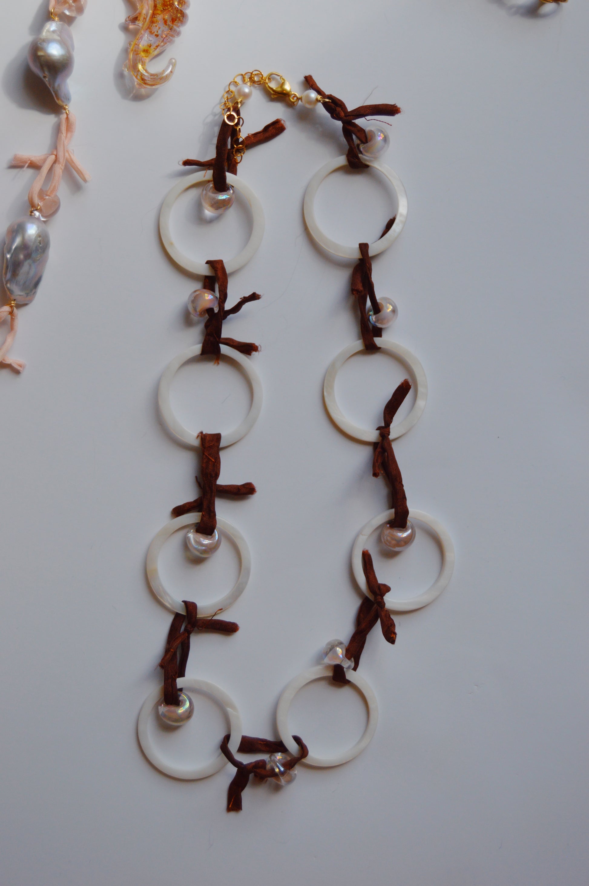 The Silk and Circle Layer is a handmade jewelry piece with large white circular links, brown leather ties, and small pearls, displayed on a white surface with glimpses of other jewelry in the corners.