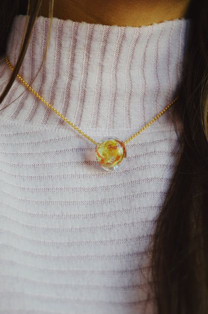 A person wears the Grand Bubble Single - Mars, a 14k gold filled chain necklace with a round, colorful glass pendant, along with a ribbed light pink sweater. Only the lower part of their face and neck are visible.