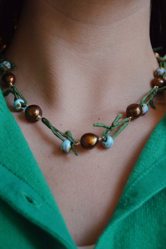 A close-up of a person wearing a green top and the Silk and Pearl Layer - Green Dunes necklace, featuring handmade glass beads with brown and blue marbled details and green fabric ties around their neck.
