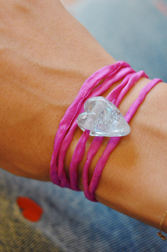 A close-up of a wrist wearing the Silk Glass Heart- Pink & Blue bracelet, featuring a handmade lampwork glass heart charm. The background displays blue denim fabric with a small red spot.