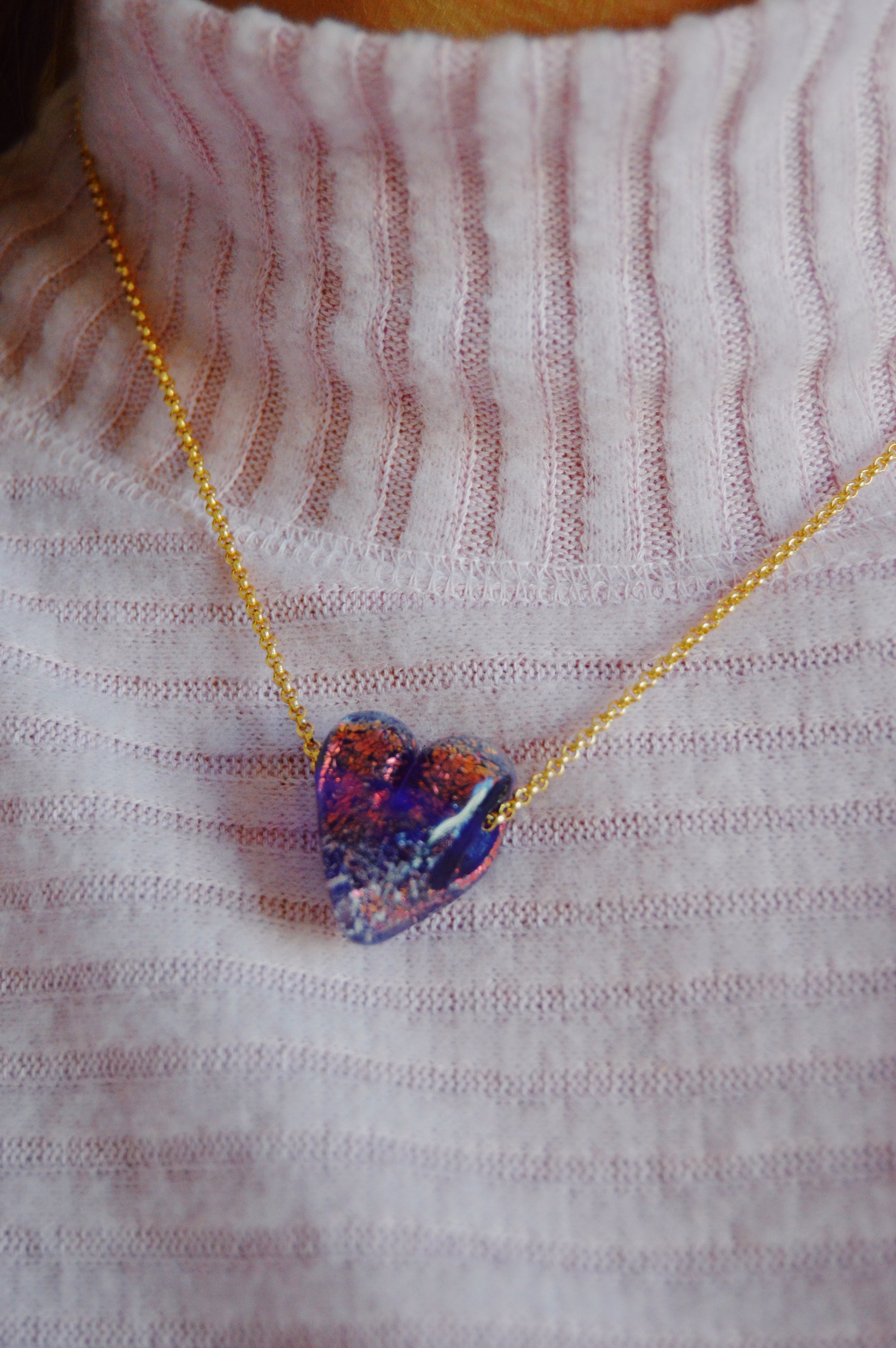 A close-up of a person wearing a ribbed light pink turtleneck sweater and the Glass Heart Necklaces- Purples, featuring an artisan gold-filled chain with a handmade purple glass heart pendant.