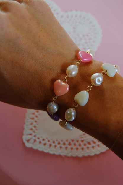 A hand wears two Candy Bracelet-White gold chain bracelets with heart-shaped and pearl beads in pink, white, purple, and cream, set against a pink background with a lace paper doily.