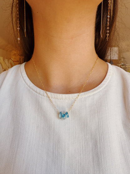 A person wears a white top, gold chain earrings, and the Dainty Donut Layer—a delicate 14K gold chain necklace with three small blue gemstones as pendants. The image highlights the neck and chest area.