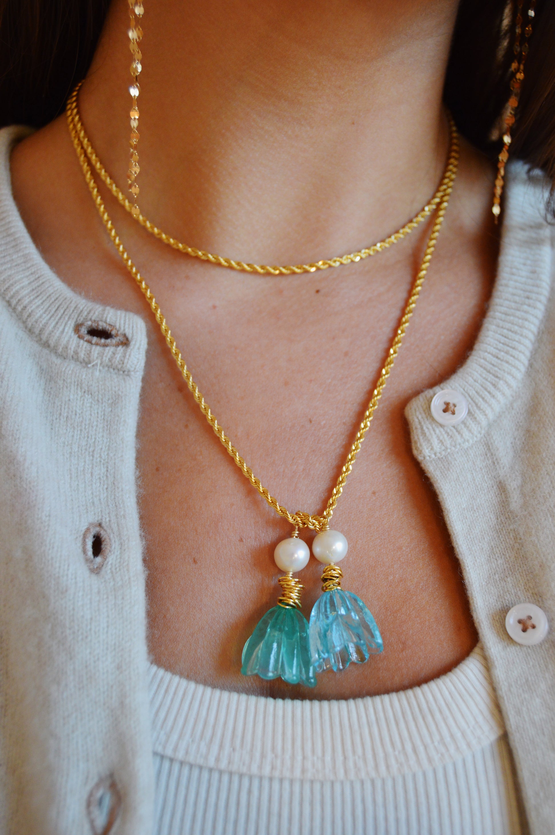 A close-up shows a person wearing the Tulip Wrap Necklace-Bluebells, featuring layered gold-filled chains with cultured freshwater pearls and blue glass lampwork tulip leaves, styled over a cream knit cardigan and white ribbed top.