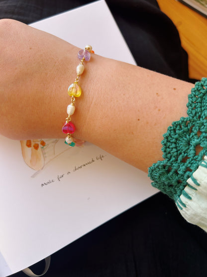 A hand wearing the Bestie Bracelet—featuring pastel acrylic hearts and pearls—rests on an open notebook with the words “make for a charmed life.” The sleeve has green crochet detailing.