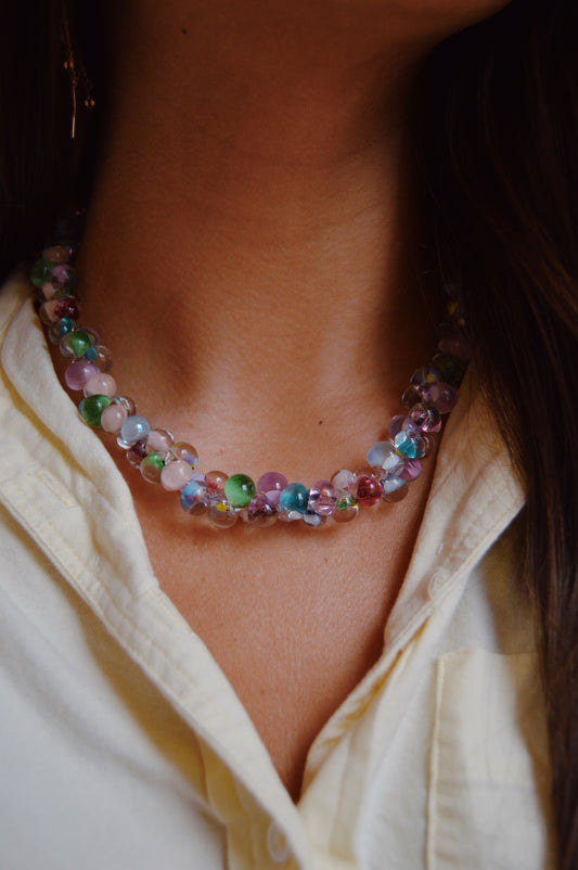 A close-up of a person wearing the Bubble Glass Necklace- Bloom Mix over a light yellow collared shirt. The necklace showcases round, translucent beads and handmade glass beads in pastel colors.