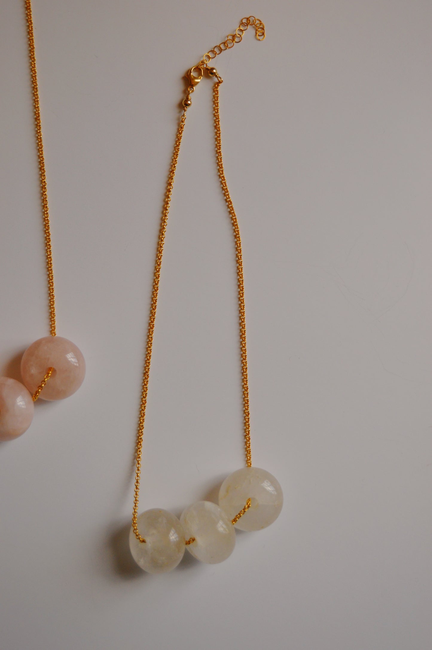 The Grand Donut Layer-Clear is a handcrafted necklace with a 14k gold-filled chain featuring three translucent round beads and a matching clasp. A similar style with pink beads appears partially on the left.