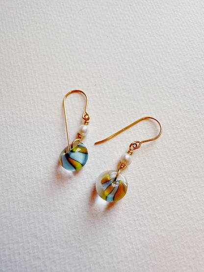 Bubble Minis are handmade earrings with gold-filled findings, each featuring a round glass bead with blue, yellow, and orange swirls topped by a freshwater pearl, shown on a textured white background.