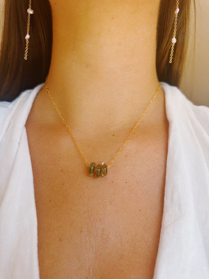 A woman in a white v-neck top wears the Dainty Donut Layer: a delicate 14K gold chain necklace with three green glass donut beads, paired with gold chain earrings featuring small pearls.