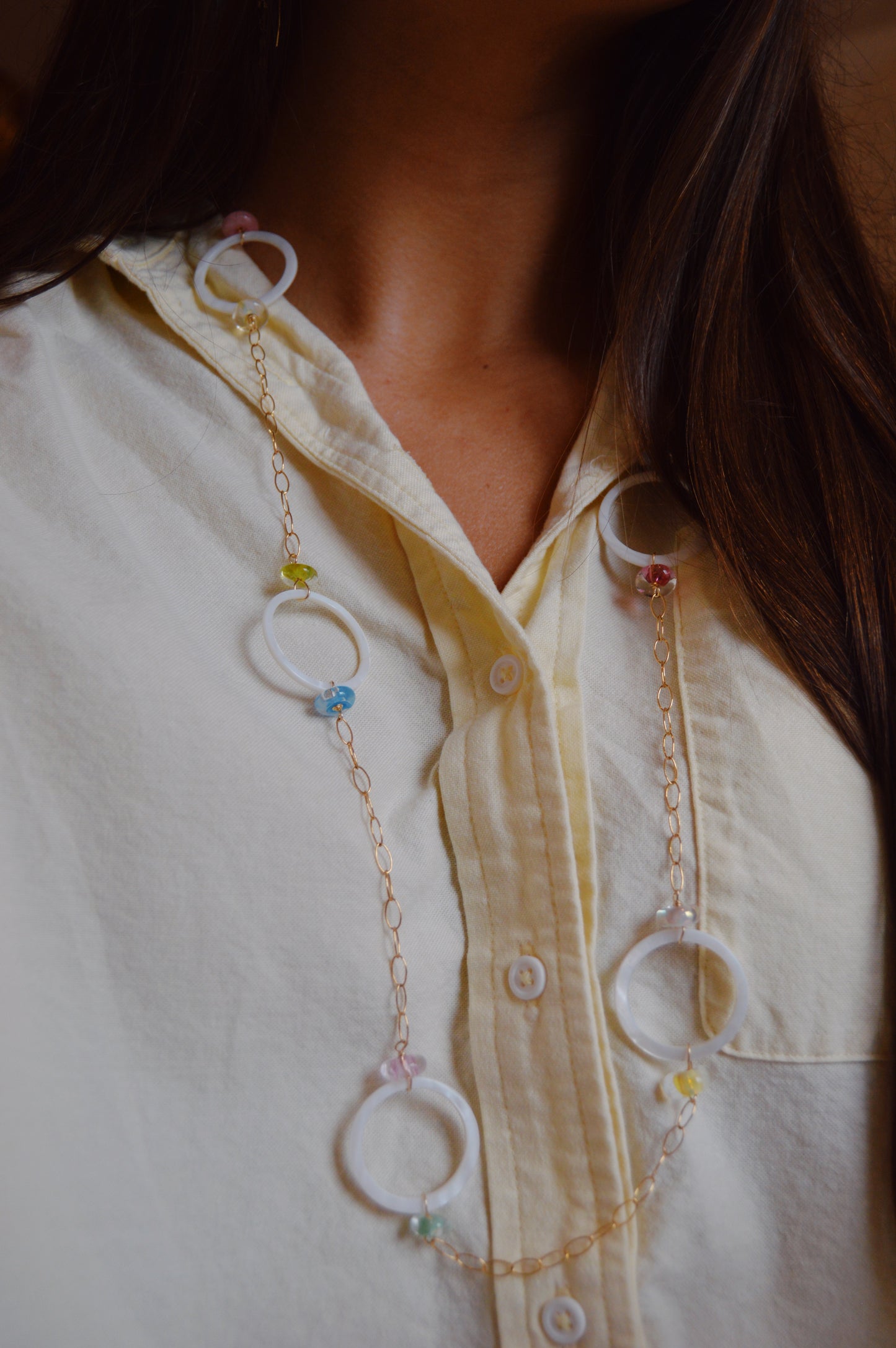A woman wears the Long Glass and Circle Layer: a light yellow button-up shirt paired with a handcrafted necklace featuring large white rings and colorful beads on a 14k gold-filled chain.