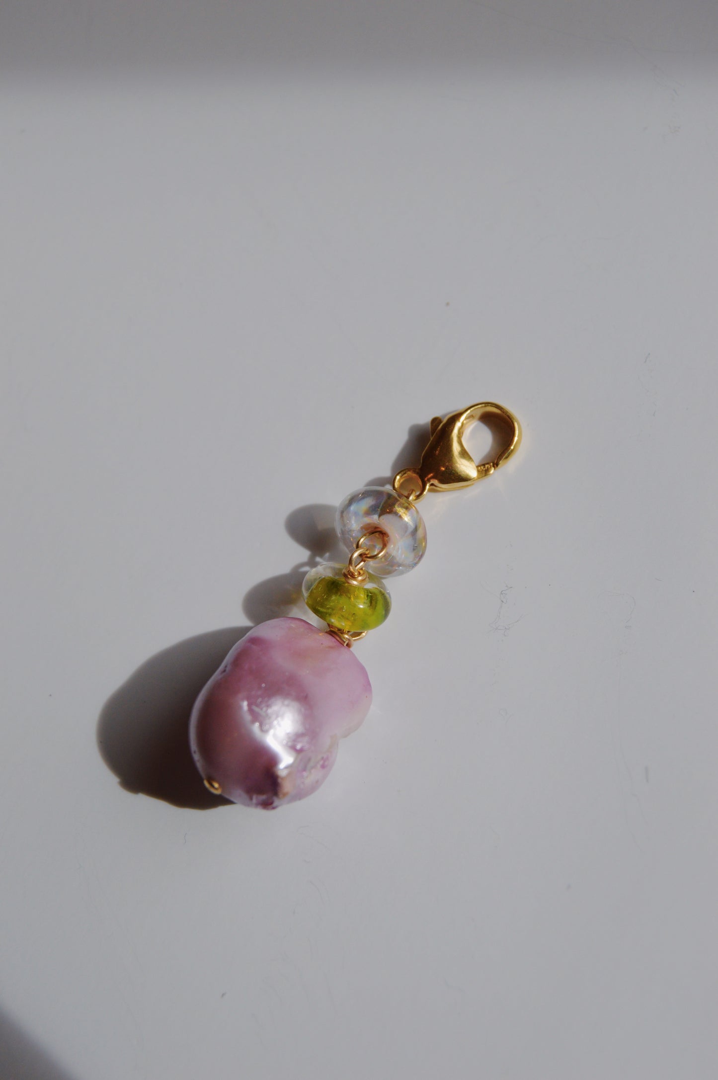 The Glass and Lavender Pearl Charm features a gold clasp, a large pink cultured freshwater pearl, a round green handmade lampwork glass bead, and a clear bead, photographed on a white surface with soft shadows.