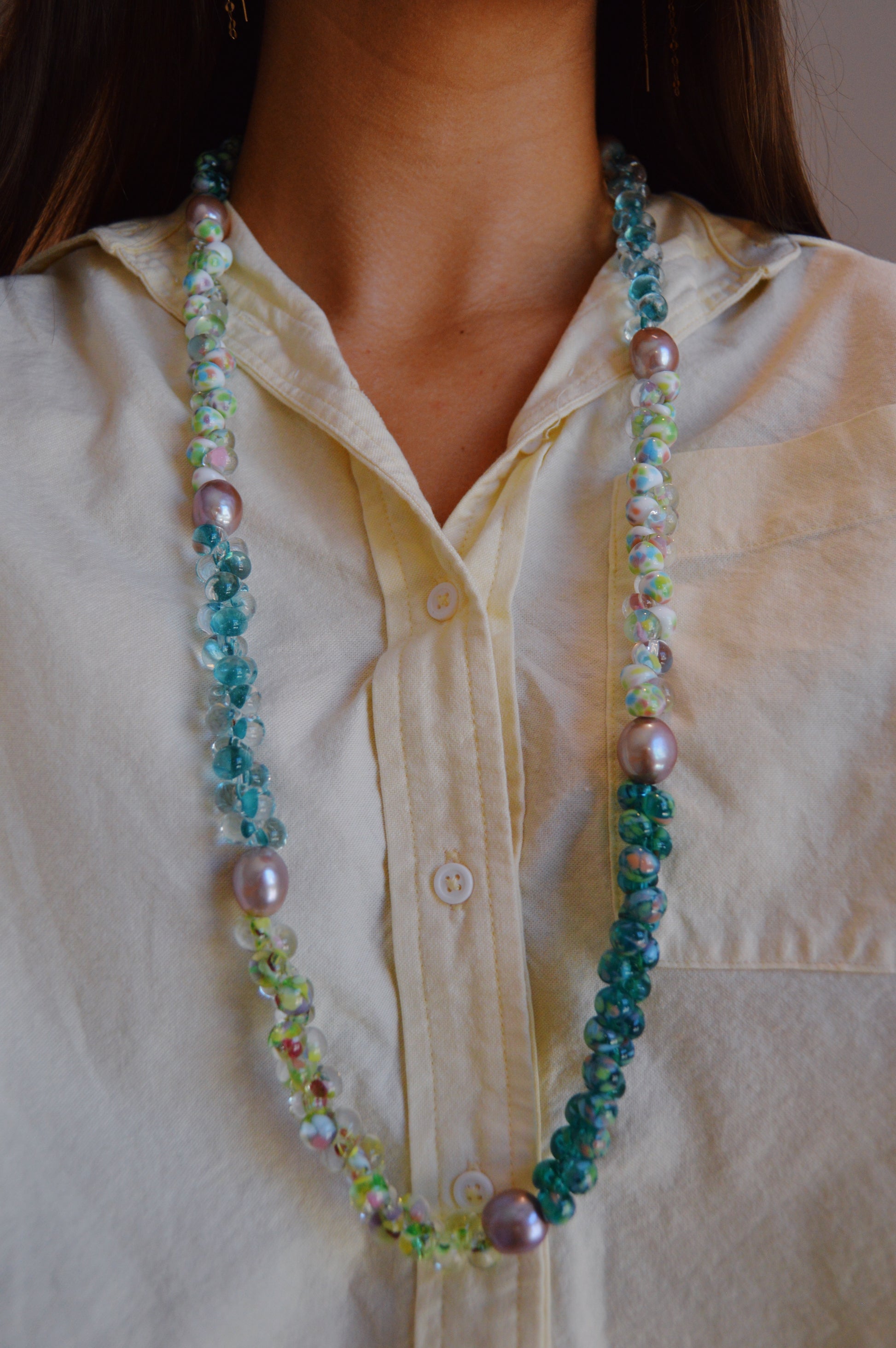 A person in a cream button-up shirt showcases the Long Bubble Glass and Pearl Necklace, featuring handmade blue, green, and iridescent lampwork glass beads accented with large cultured freshwater pearls.