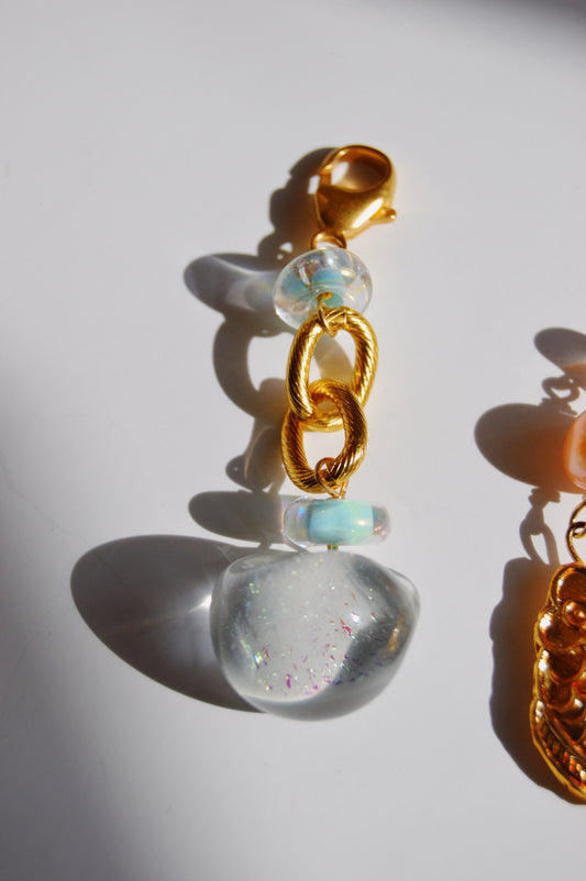 The Grand Bubble Charm- Light Blue features two twisted 14k gold-filled links, handmade lampwork glass beads, and a clear resin pendant with sparkles, casting a shadow on a white surface.