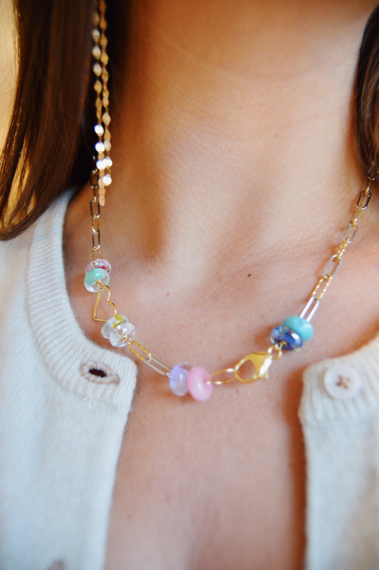 A close-up of a person wearing the Valentine Clasp in Front Heart Necklace, featuring a gold chain with colorful pastel glass beads, paired with a light-colored cardigan with button details.