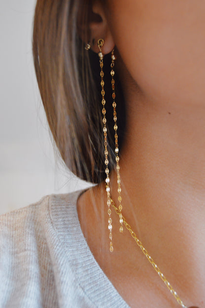 Close-up of a person wearing Sway Studs—long, delicate 14k gold filled earrings—and a matching gold necklace. The person has straight brown hair and is dressed in a light gray top.