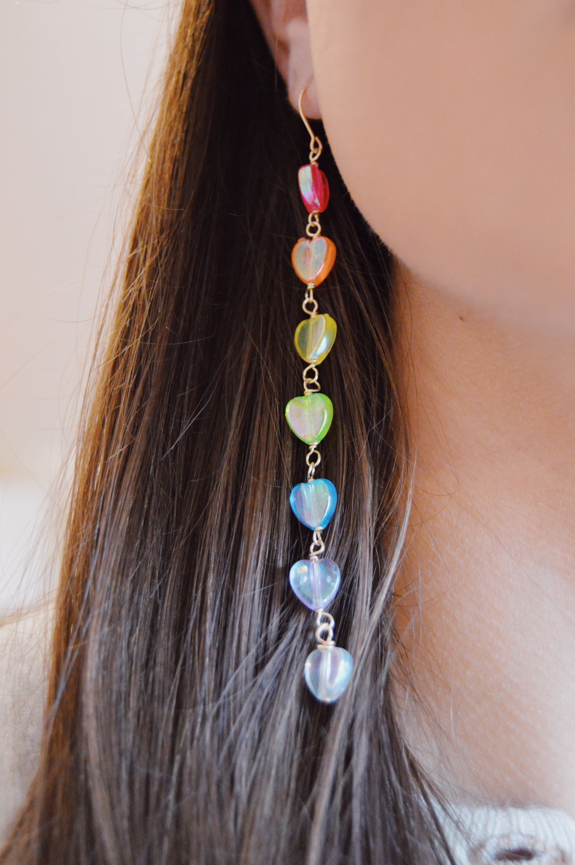 A close-up of a person wearing Valentine Rainbow Acrylic Drops, featuring seven vertically arranged colorful acrylic heart beads and 14k gold filled findings, dangling from their straight brown hair.