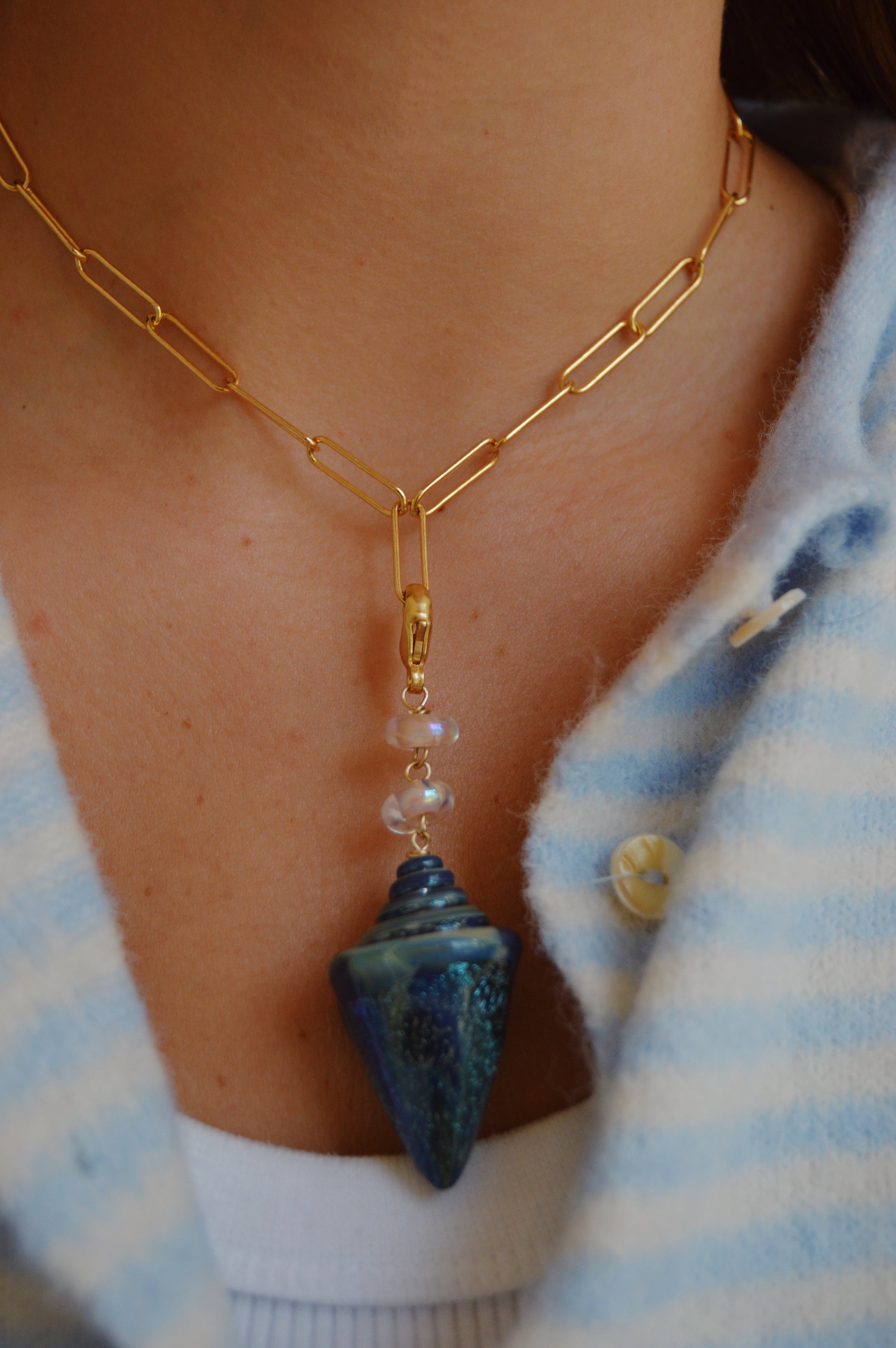 A close-up of a person wearing the Glass Shell Charm, a 14k gold-filled chain necklace featuring a blue, cone-shaped pendant with lampwork glass beads and clear accents, paired with a blue and white striped fuzzy sweater and white top.