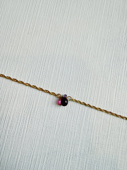 The Dewdrop Bracelet features a delicate 14k gold filled chain with a small, teardrop-shaped purple pendant resting on a textured white surface.