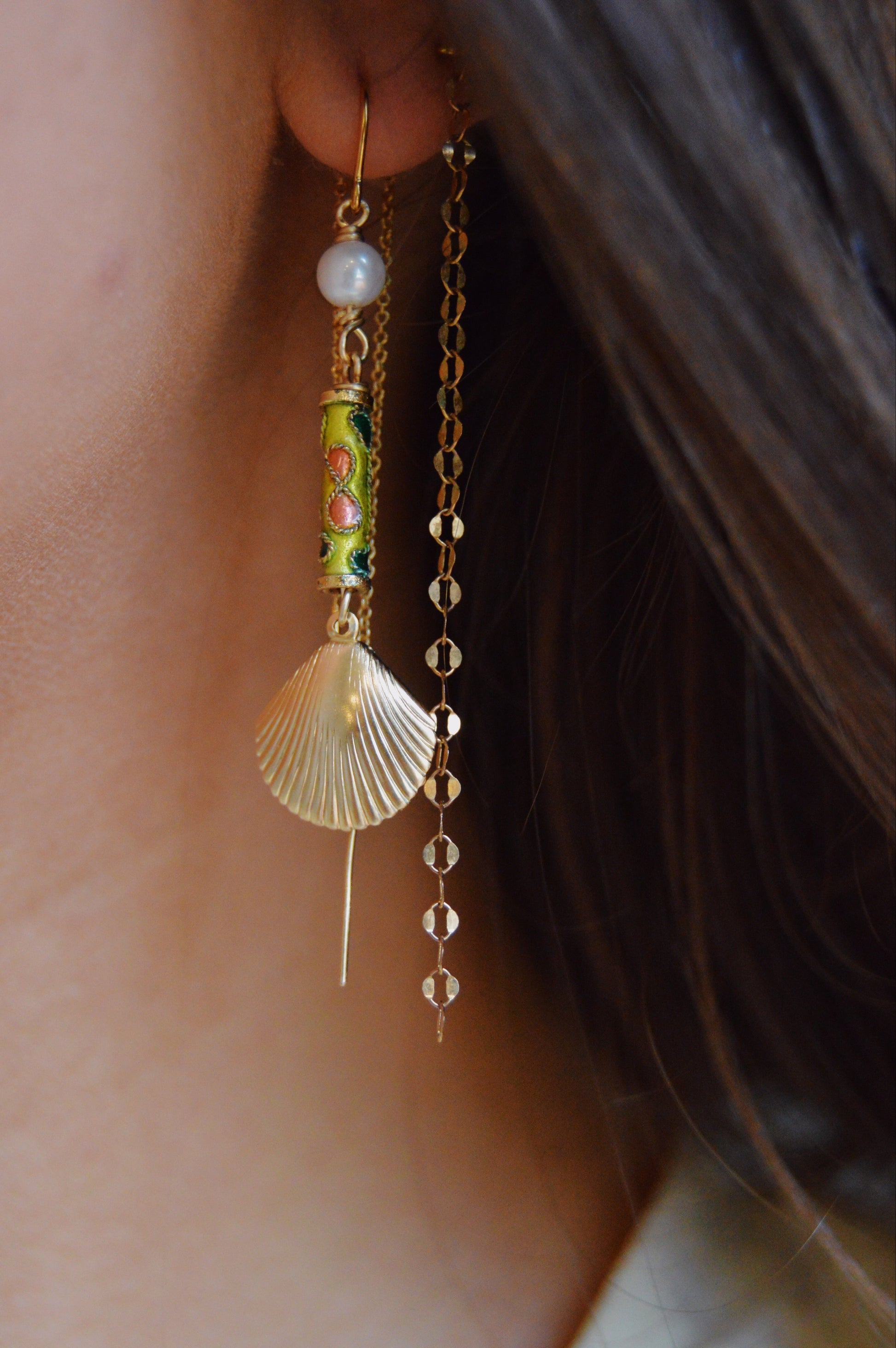 A close-up of a person's ear wearing Cloisonne Shell Threaders: decorative earrings featuring cultured freshwater pearls, a green and gold cylindrical bead, a gold seashell, and a long 14k gold filled chain. Brown hair frames the ear.