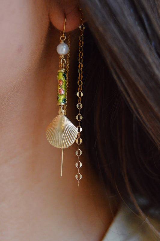 A close-up of a person's ear wearing Cloisonne Shell Threaders: decorative earrings featuring cultured freshwater pearls, a green and gold cylindrical bead, a gold seashell, and a long 14k gold filled chain. Brown hair frames the ear.