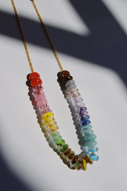 The Rainbow Lots of Donuts necklace features handblown glass beads in a rainbow gradient from red to blue on a gold chain, casting soft shadows. Finished with gold filled findings for an elegant touch.