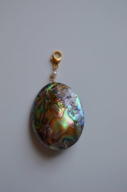 The Abalone Charm features a round abalone shell with an iridescent finish, accented by a gold-filled lobster clasp and a small freshwater pearl, shown on a plain light background.