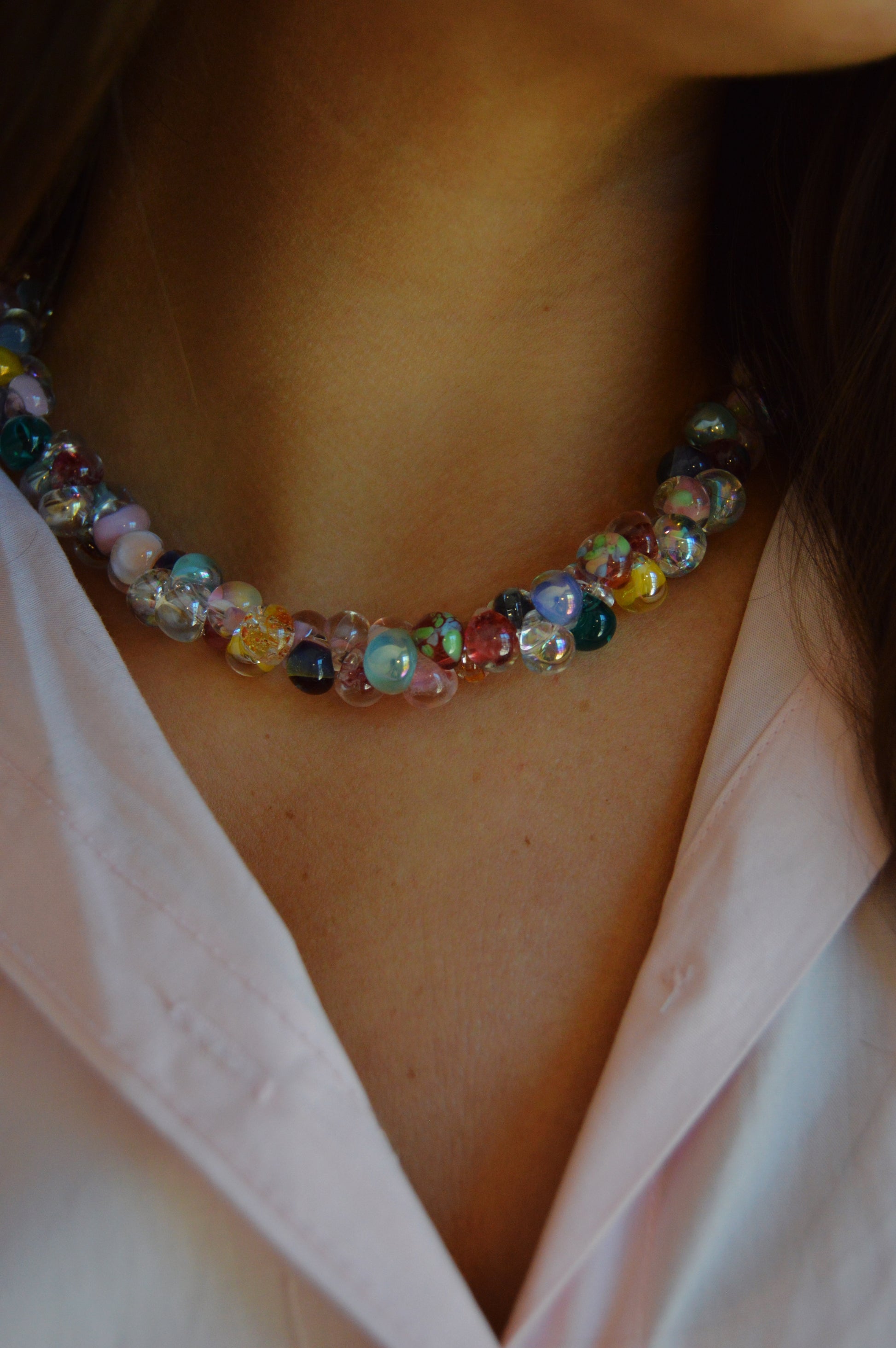 A close-up of a person wearing a pastel pink collared shirt and the Valentine Mix Bubble Glass Necklace, featuring round beads in blue, yellow, red, and clear. The person's face is partially out of frame.
