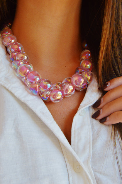 A person wearing a white collared shirt styles the Grand Bubble Glass Necklace-Shimmer Pink, featuring chunky iridescent pink beads. One hand with brown-painted nails lightly touches the shirt near the collar.