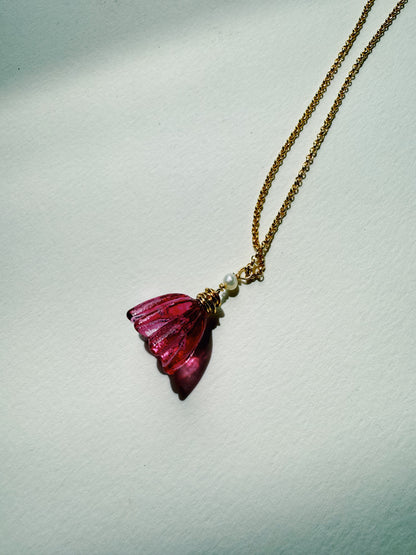 The Tulip Necklace, featuring a handblown glass tulip pendant in translucent pink and accented with two small cultured freshwater pearls, is displayed on a light, textured surface in bright natural light.