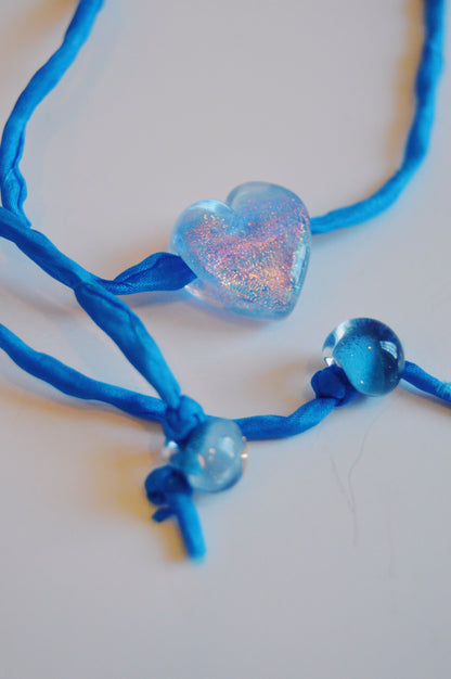 A close-up of the Silk Glass Heart - Cobalt necklace, featuring a handmade lampwork glass heart and artisan craftsmanship on blue silk fabric, displayed against a white background.