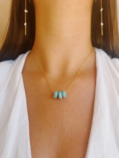A woman wears the Dainty Donut Layer, a delicate 14K gold chain necklace with four turquoise beads and a gold filled clasp, paired with gold drop earrings with small pearls. She is dressed in a white V-neck top.