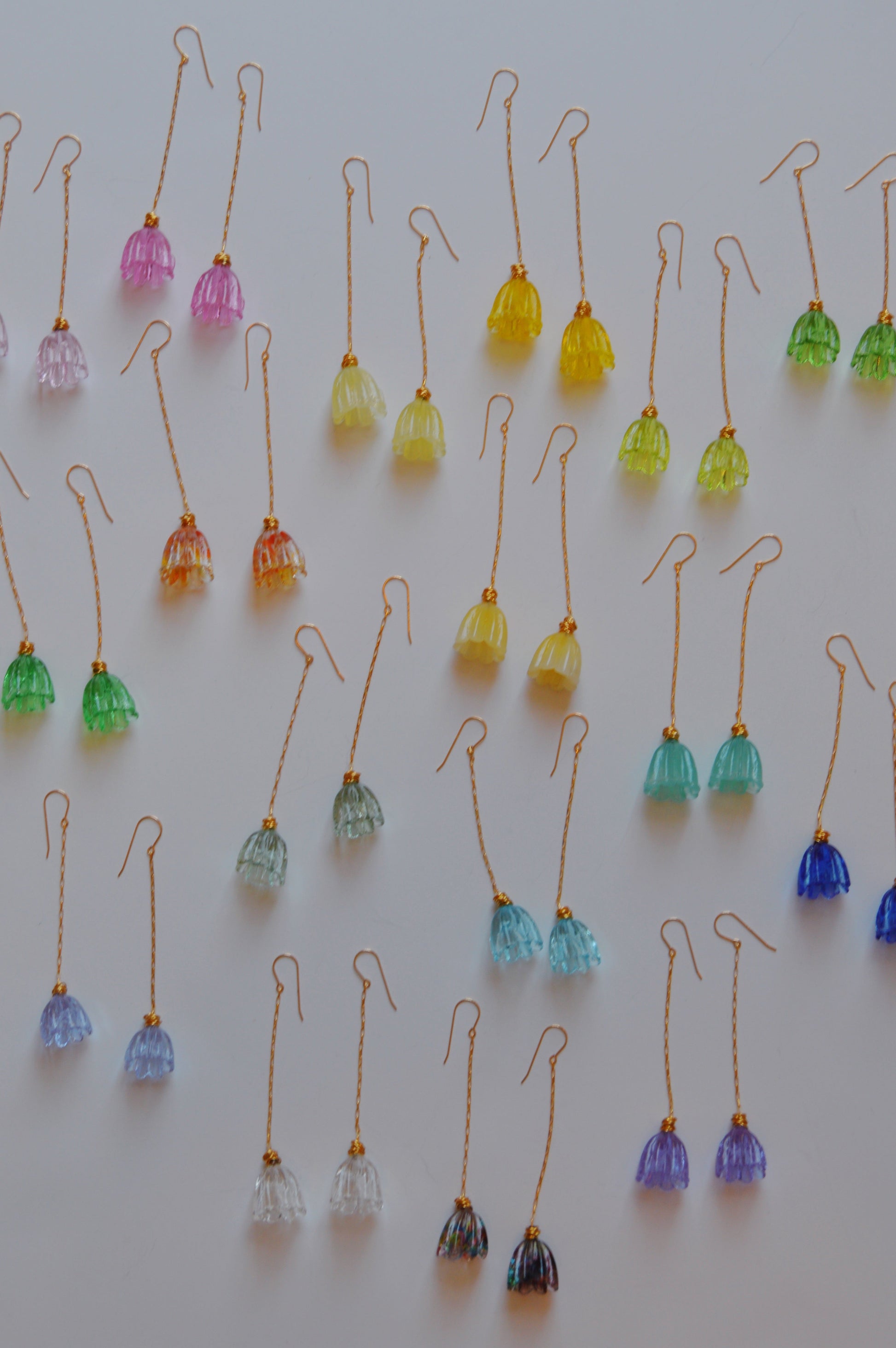 Glass Tulip Earrings, handmade and shaped like small bell flowers, are arranged in neat rows on a white surface. Available in pink, yellow, green, blue, and clear—ideal for glass artists and enthusiasts.
