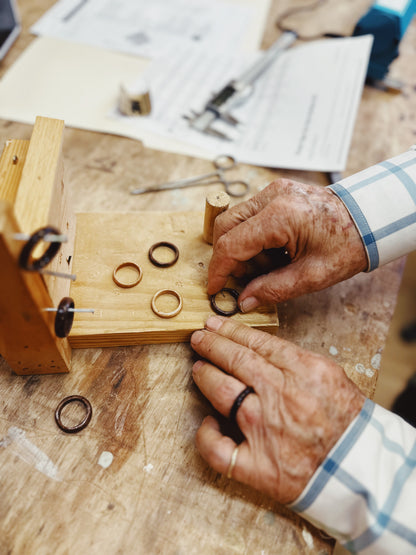 An older person’s hands work with Wooden Ring- Striped white oak wooden rings on a jig at a workbench, surrounded by papers, tools, and ring parts that highlight the artisan craftsmanship involved in making these unique striped rings.