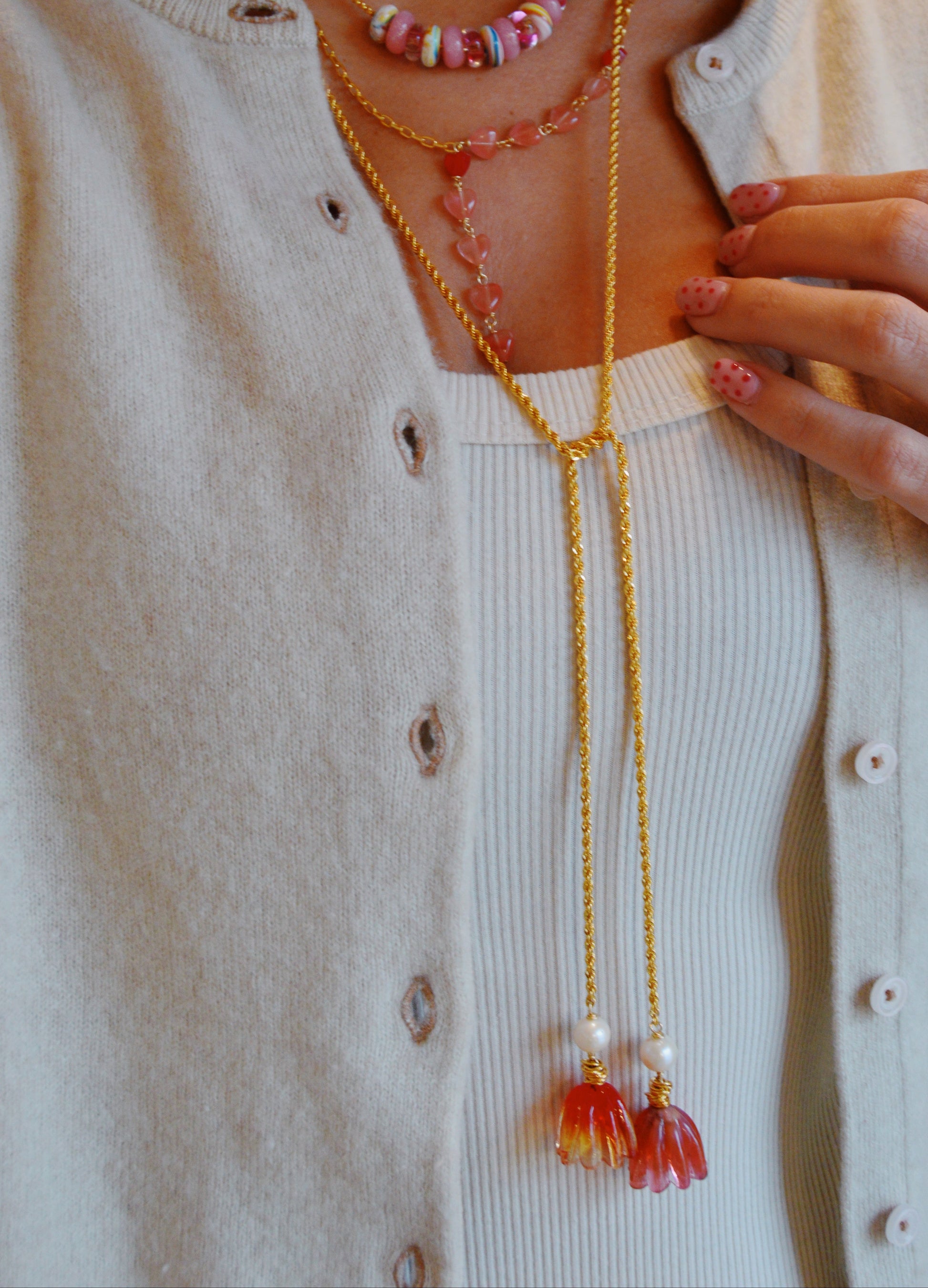 A person wearing a cream cardigan, white ribbed top, pink polka-dot nails, and the Tulip Wrap Necklace-Cherry on Top—a gold layered necklace with colorful beads and floral pendants.