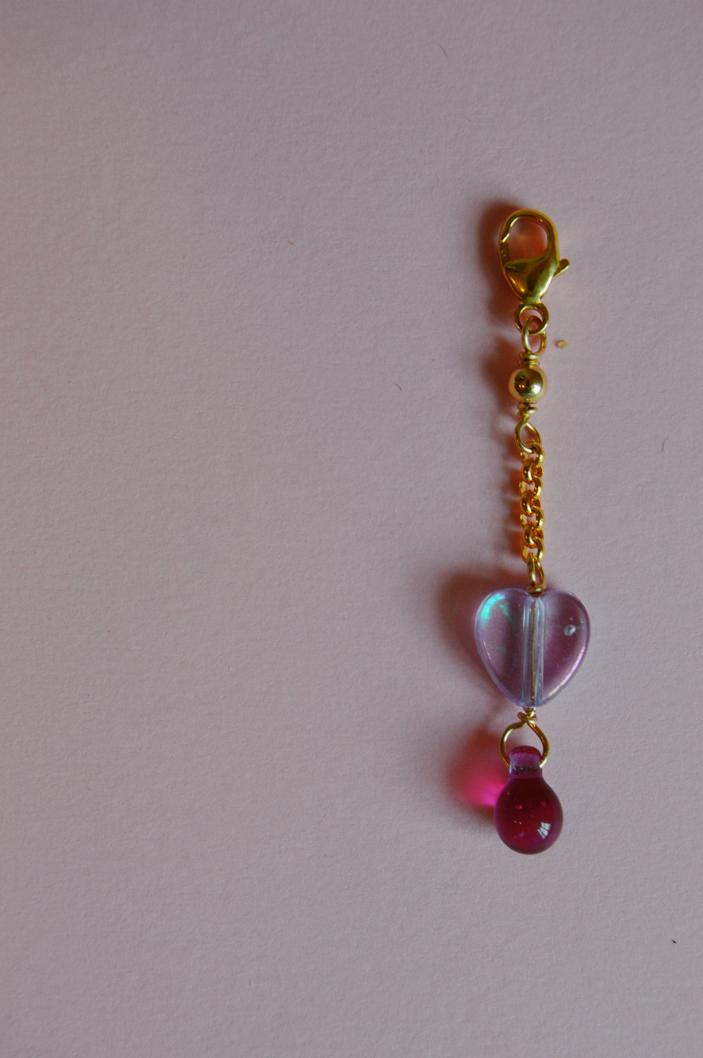 The Sweetheart Charm keychain, with a short gold chain, features a translucent purple heart-shaped charm and a small pink bead, displayed on a pale pink background.