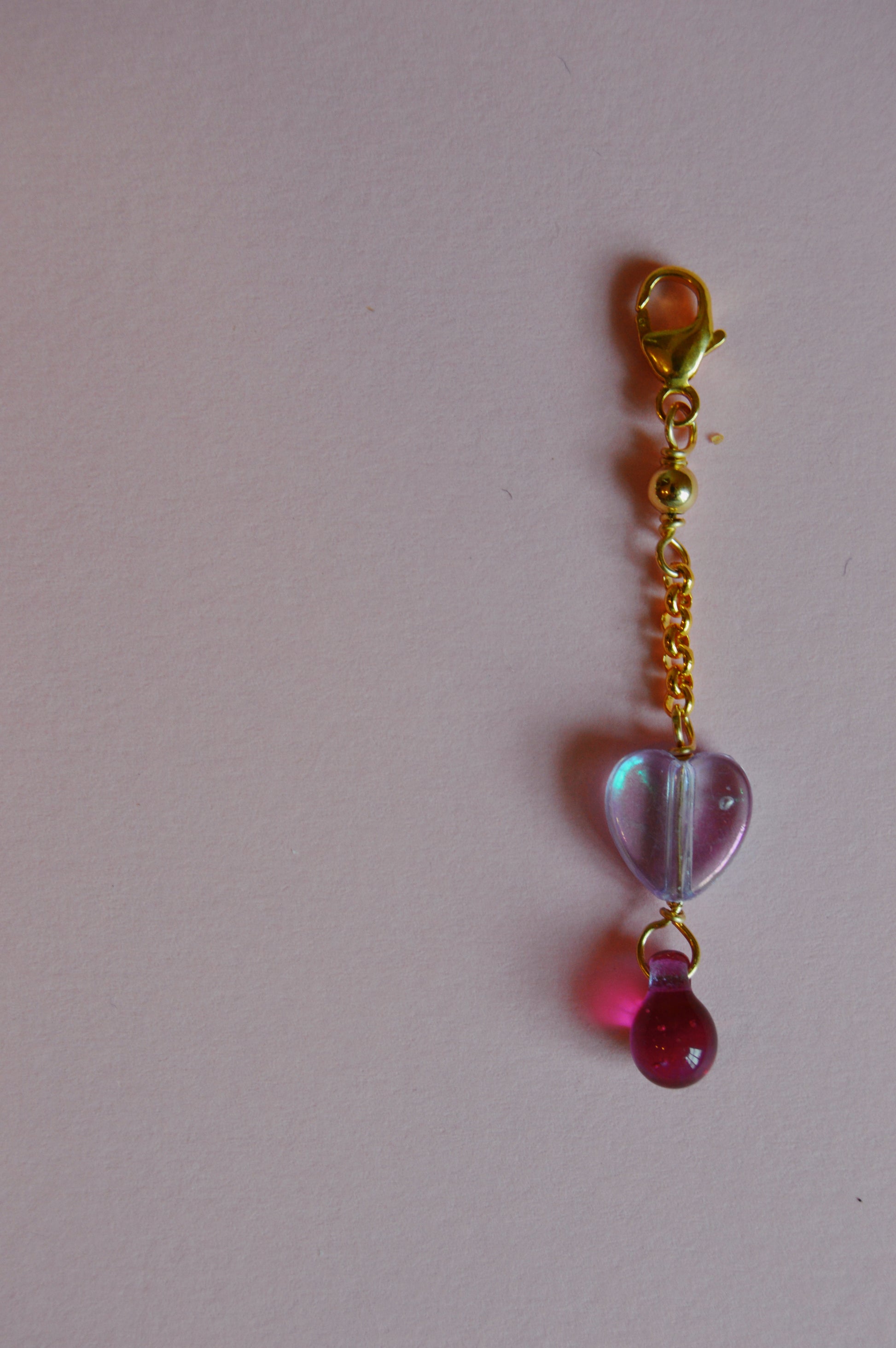 The Sweetheart Charm keychain, with a short gold chain, features a translucent purple heart-shaped charm and a small pink bead, displayed on a pale pink background.