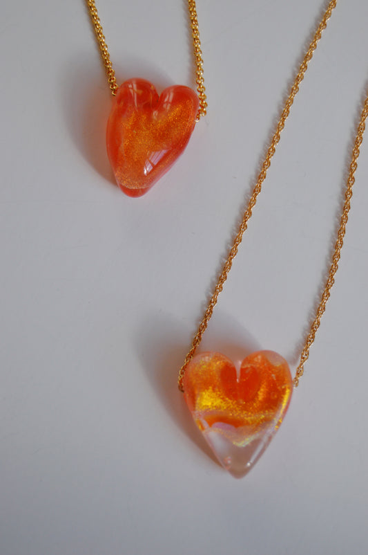 Glass Heart Necklaces- Reds & Oranges: Two gold-filled chains feature artisan heart pendants—one translucent orange handmade lampwork glass, the other orange and clear with gold accents—displayed on a plain white background.