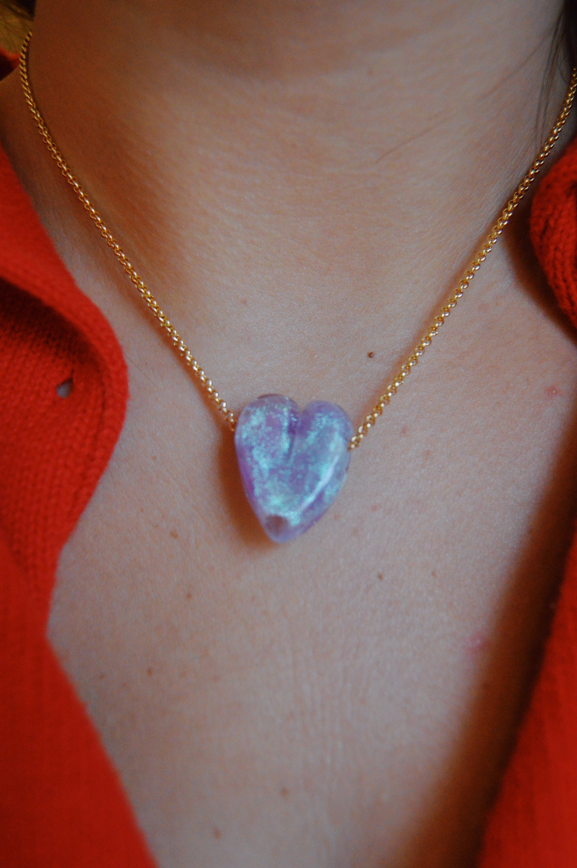 A close-up of a person wearing the Glass Heart Necklaces- Purples, featuring a gold-filled chain and a light purple handmade lampwork glass heart pendant, set against their skin and a red garment.