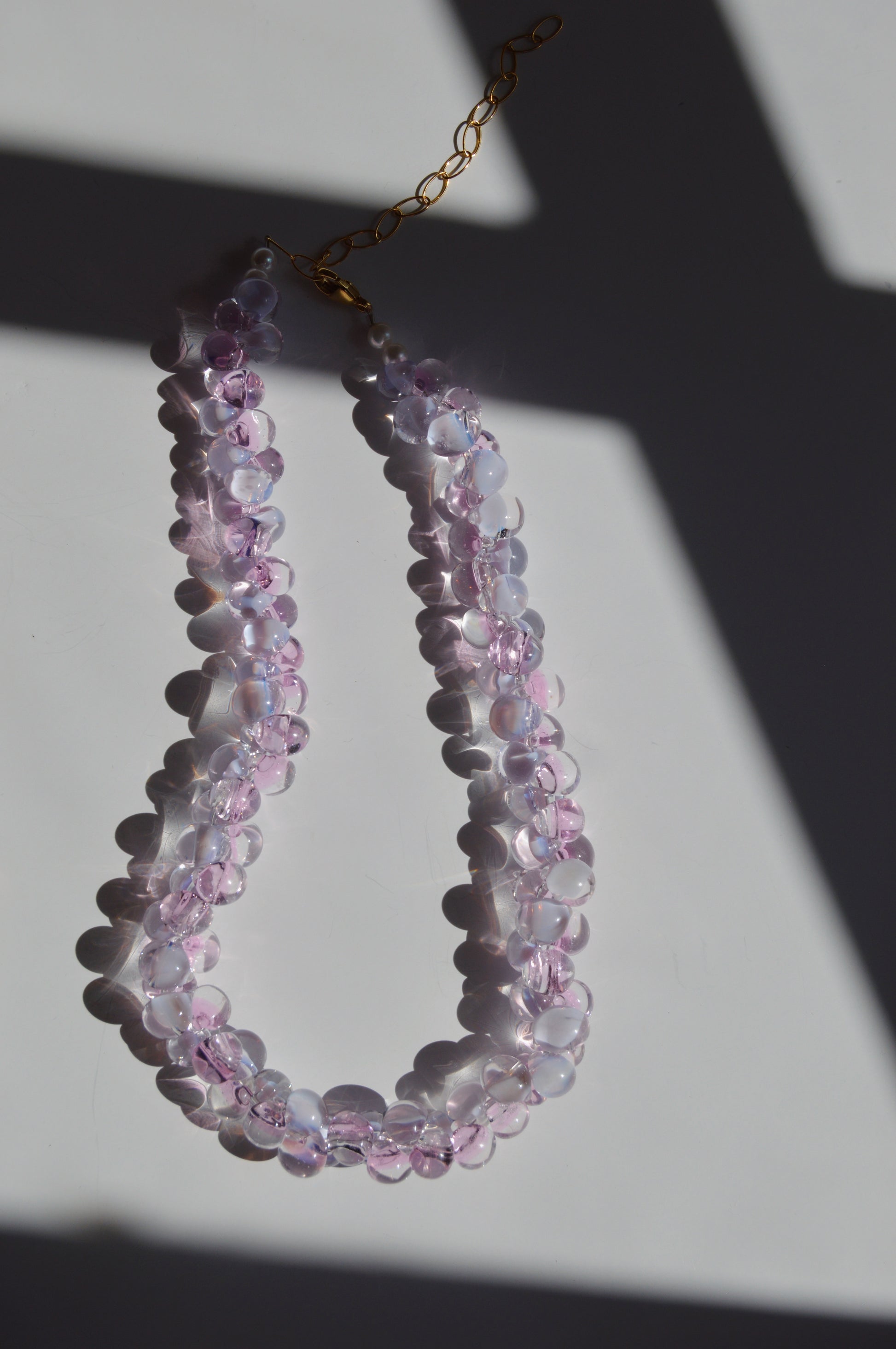 The Bubble Glass Necklace- Pink and Lavender, a double-strand handmade piece featuring translucent pink and lavender beads, is displayed on a white surface with dramatic shadows and sunlight highlighting its gold-filled findings.
