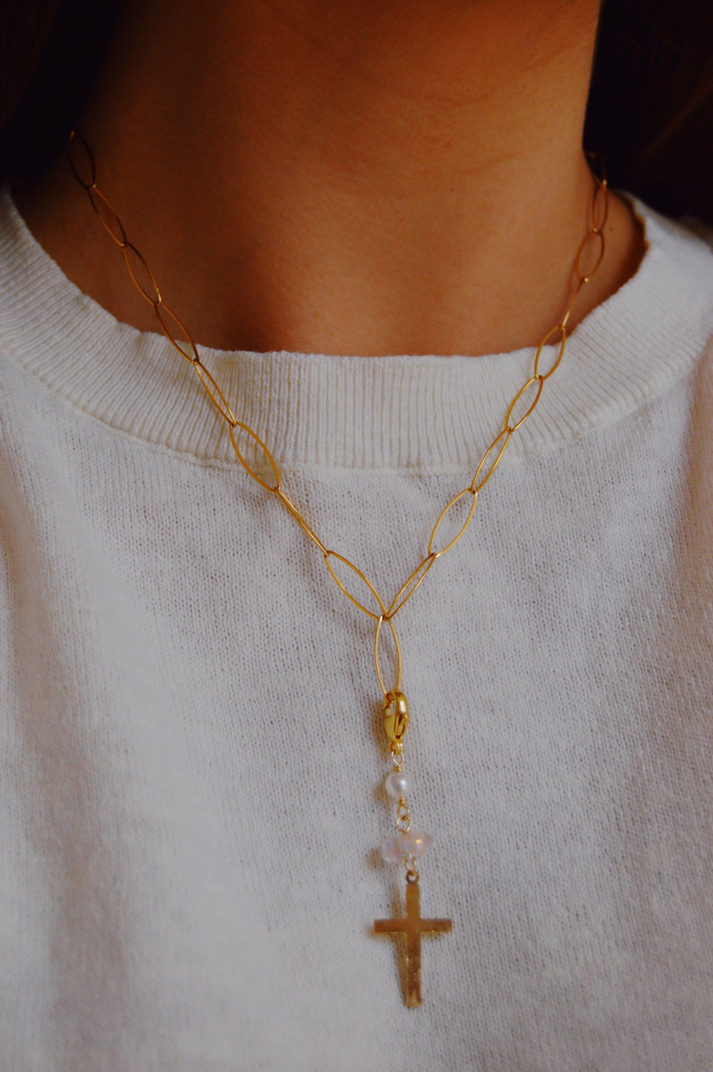 The Glass and Cross Charm features a gold-filled chain with elongated links and a 14k gold filled cross pendant, styled over a white knit top and shown on the model’s neck and upper chest.