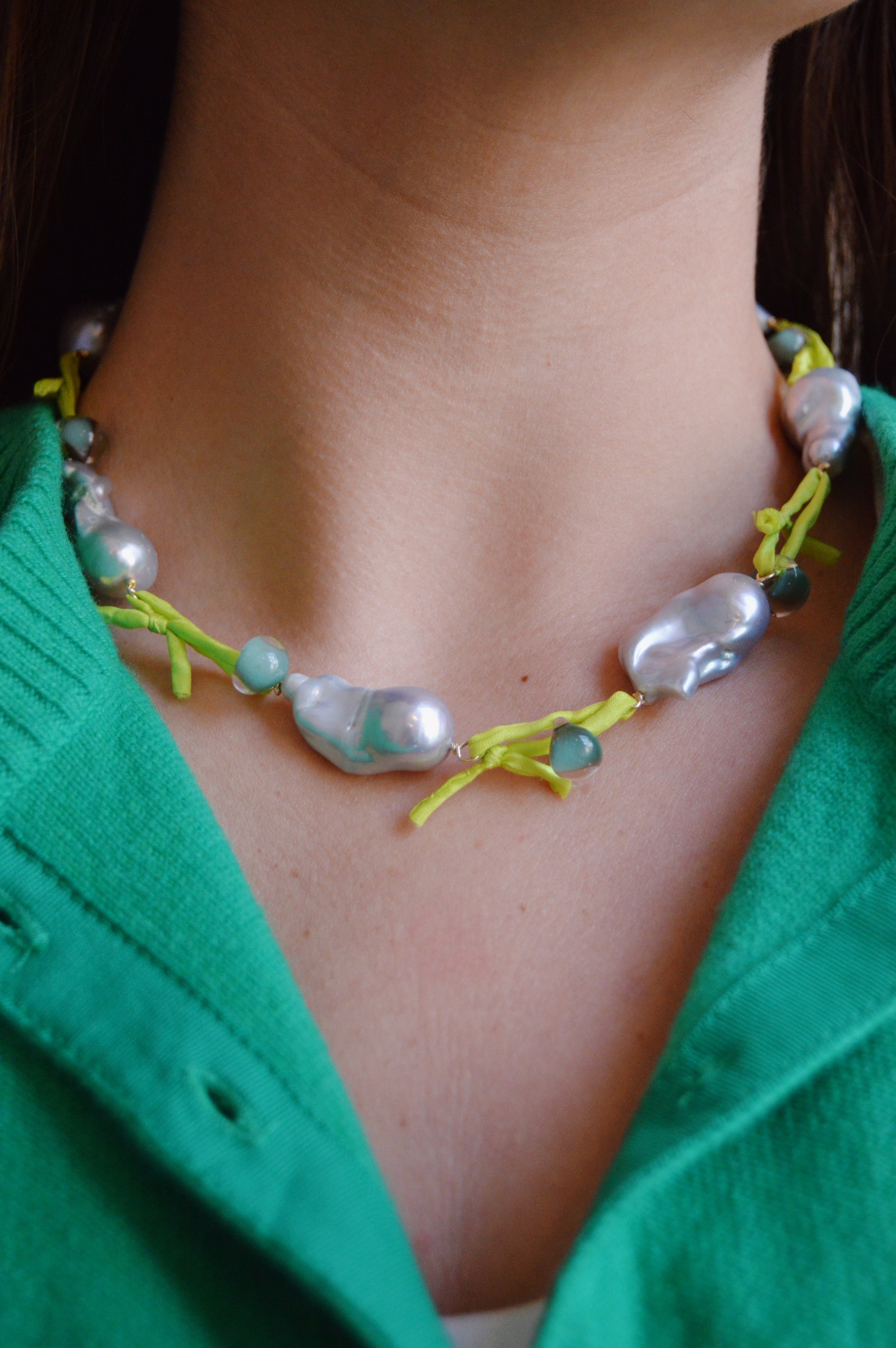 A close-up of a person wearing the Silk and Pearl Layer- Seafoam & Lime necklace—handmade with large irregular freshwater pearls, round beads, and yellow string—paired stylishly with a green cardigan.